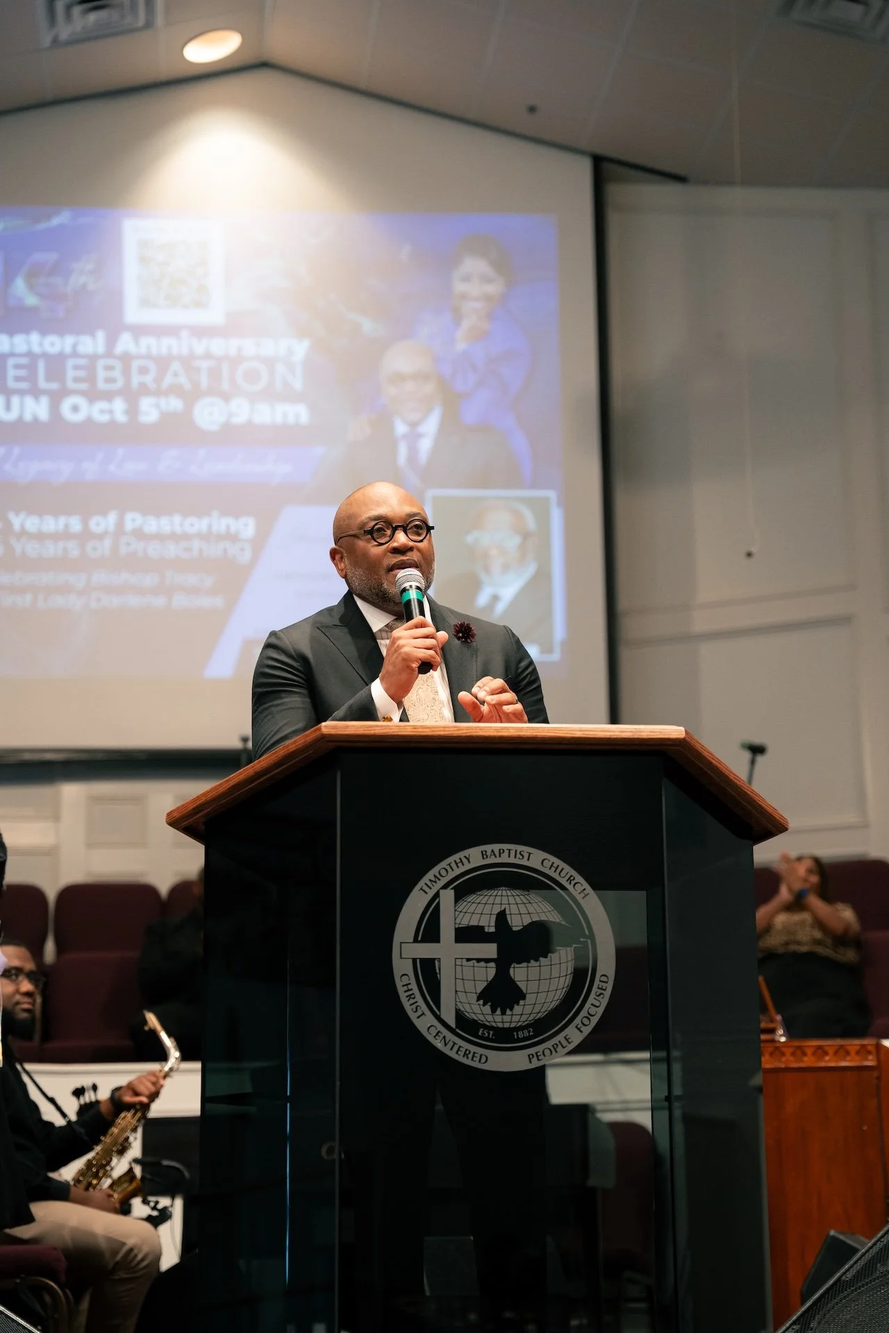 A man in a suit and glasses speaking at a church podium with the Timothy Baptist Church logo in front. In the background, there is a large projected image celebrating a pastoral anniversary event.
