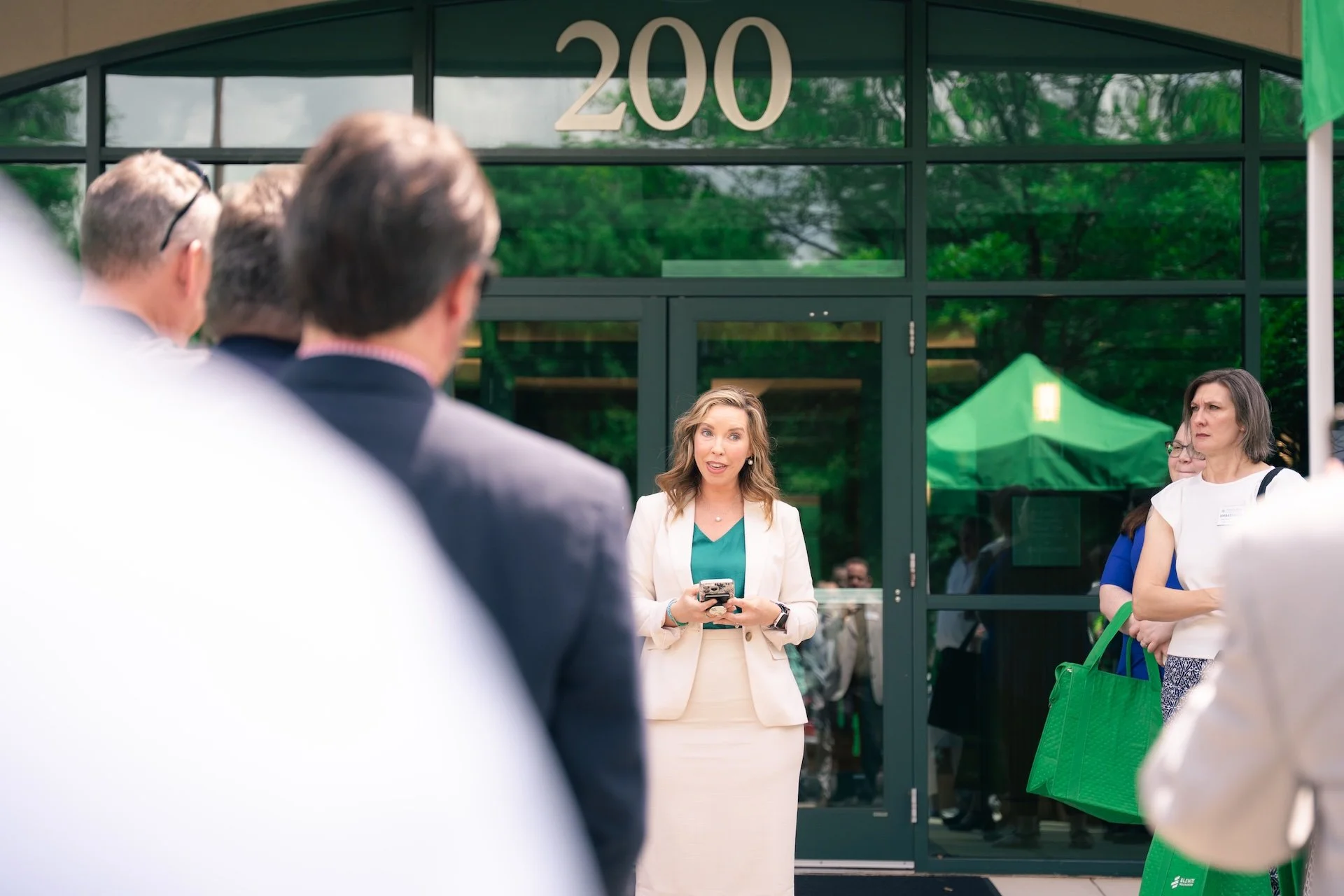 A woman in a white blazer and teal blouse speaking to a group outside a building with the number 200 on the glass door.
