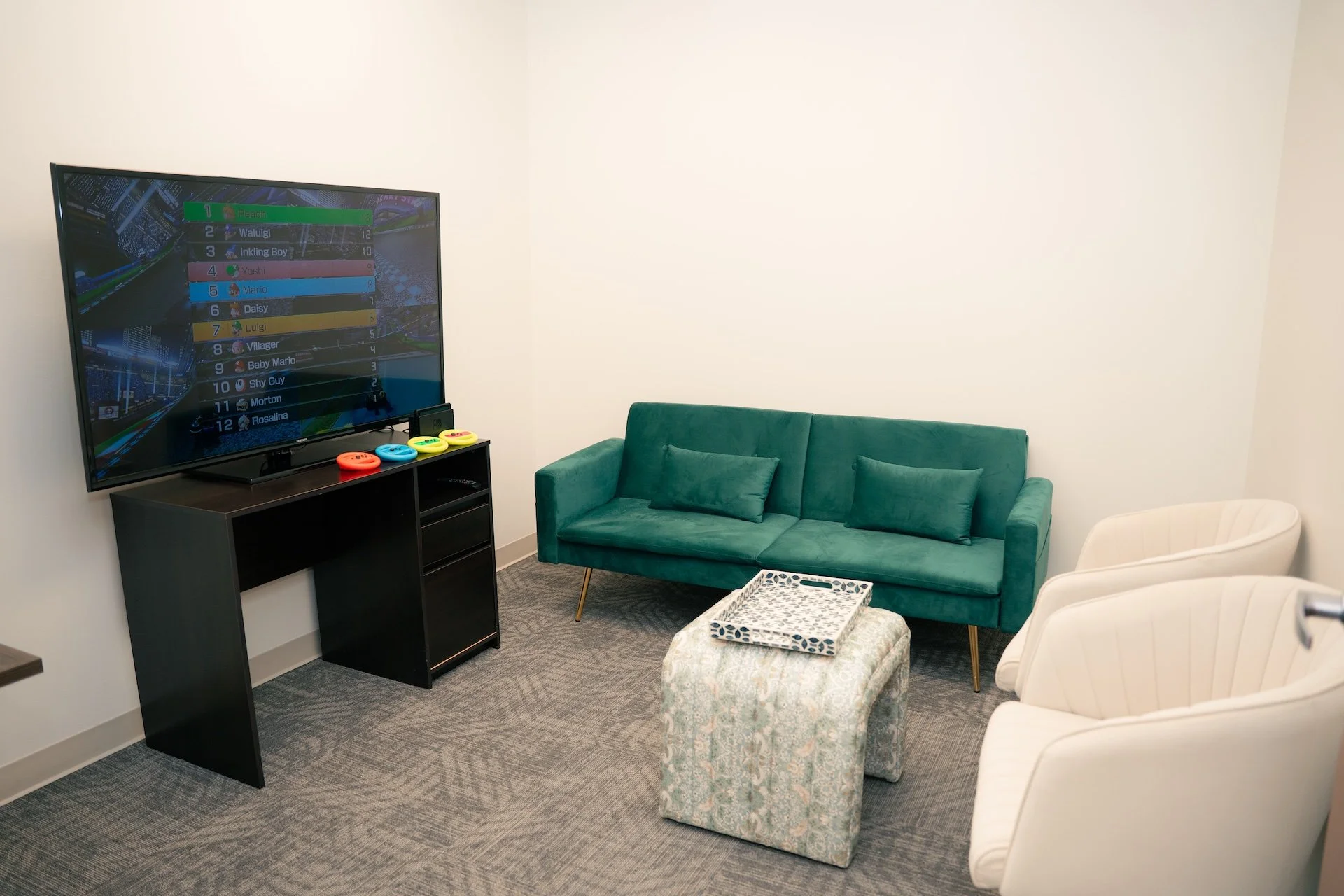 A cozy living room with a flat-screen TV displaying a racing game, a teal sofa with matching pillows, two white armchairs, a patterned ottoman with a tray, and a black TV stand with color-coded controllers.