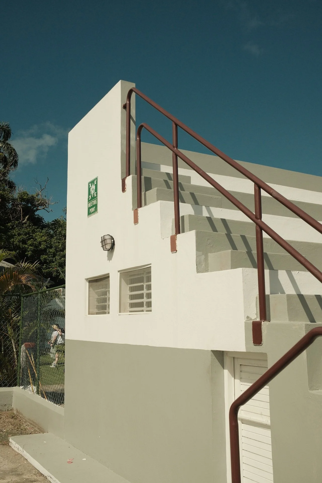 White building with exterior staircase, brown handrails, a green 'Meeting Point' sign, and a wall-mounted light fixture, set against a clear blue sky.
