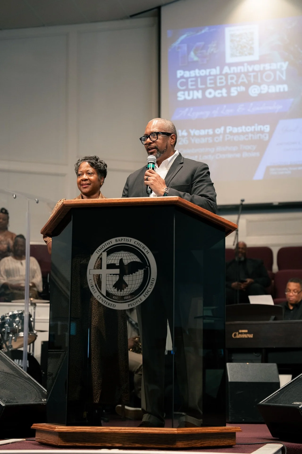 A speaker at a church podium giving a speech during a church anniversary celebration, with a woman standing beside him and a band playing in the background.