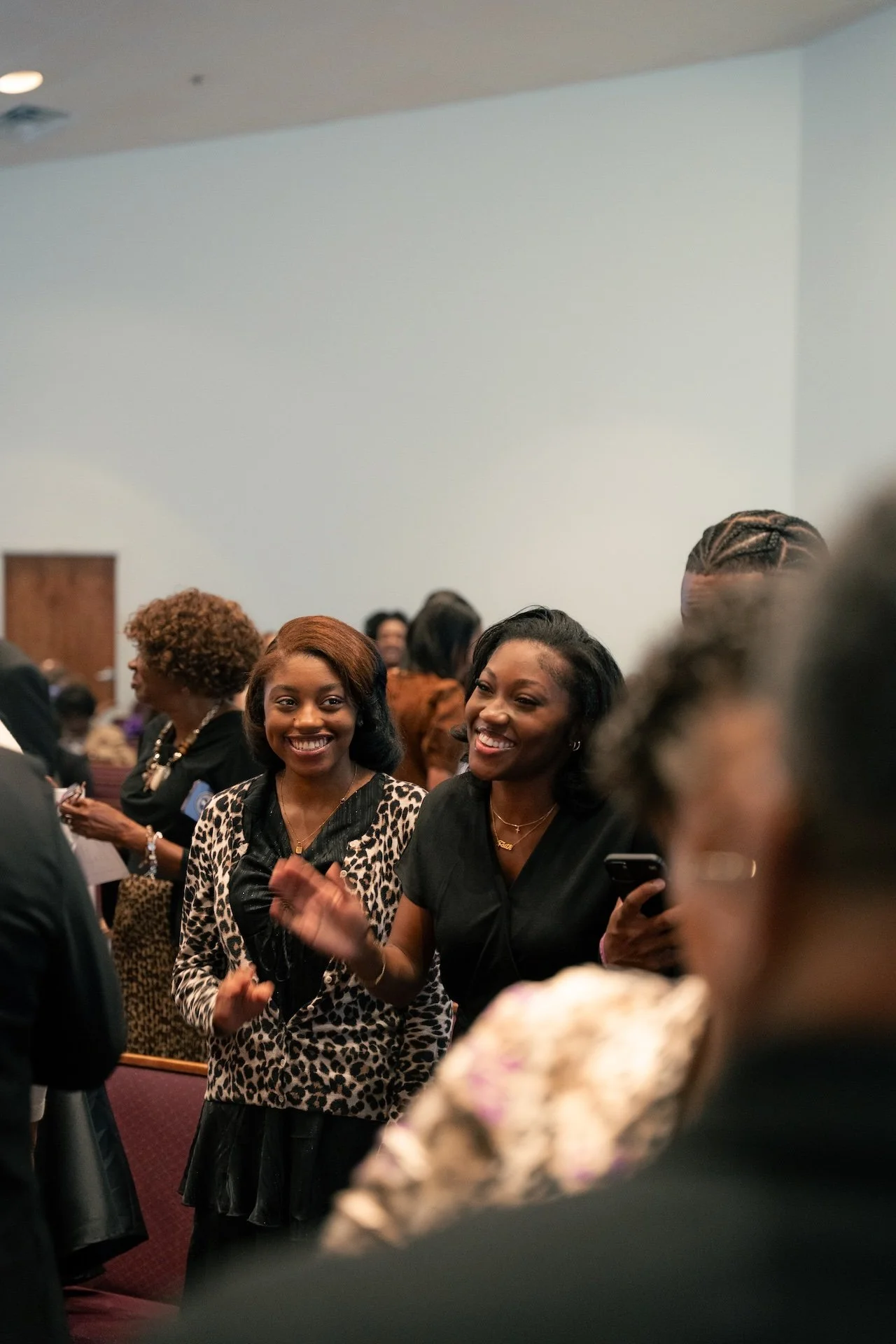 Two women smiling and talking at a social gathering, with several other people in the background in an indoor setting.