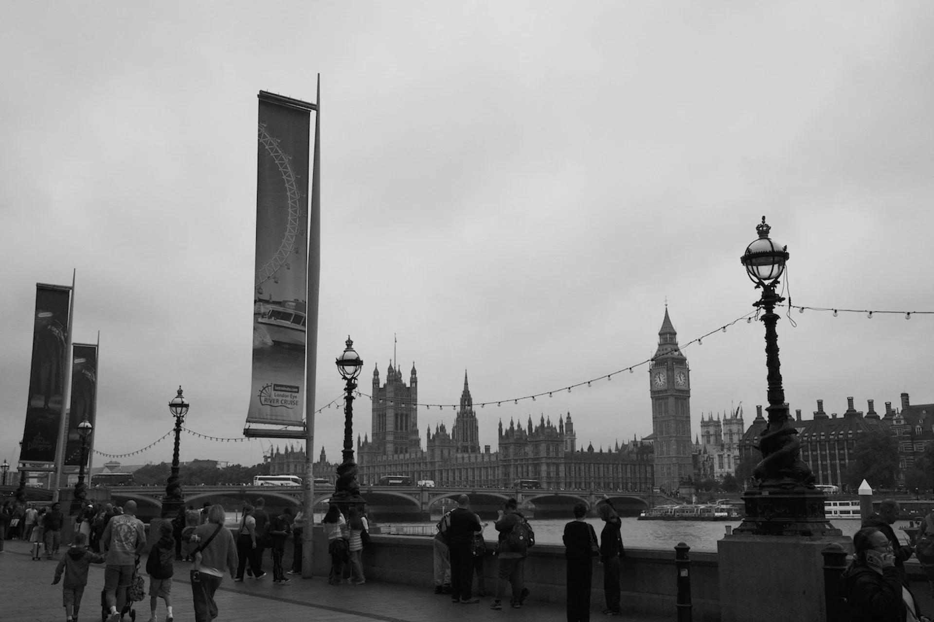 Black and white photo of London with people walking along the Thames River, Big Ben and the Houses of Parliament in the background, and a London Eye ferris wheel reflected in a vertical banner on the left.