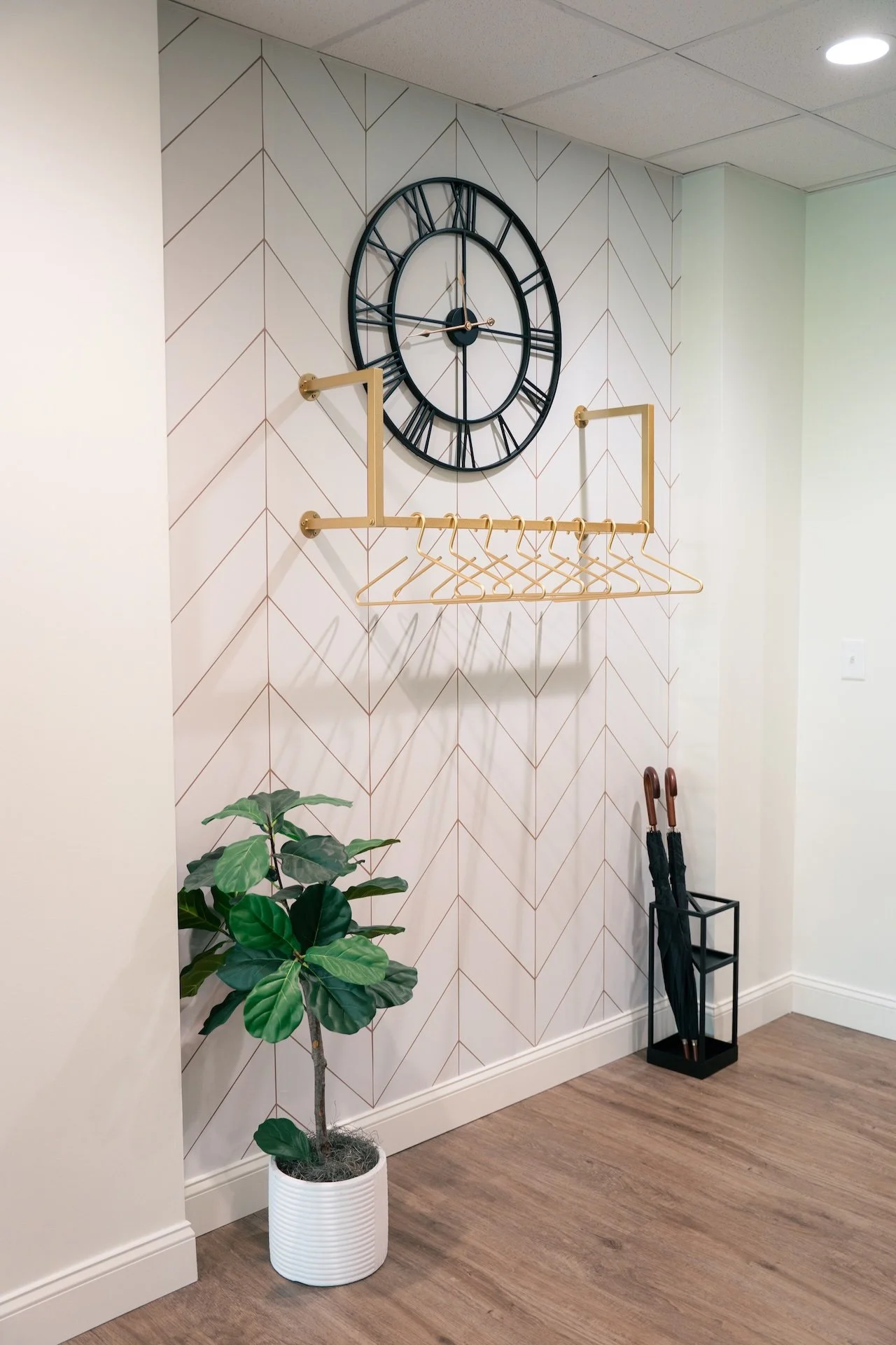A wall clock with Roman numerals, a coat rack with hangers, a potted plant, and umbrellas in a stand are in a room with wood flooring and geometric patterned wallpaper.