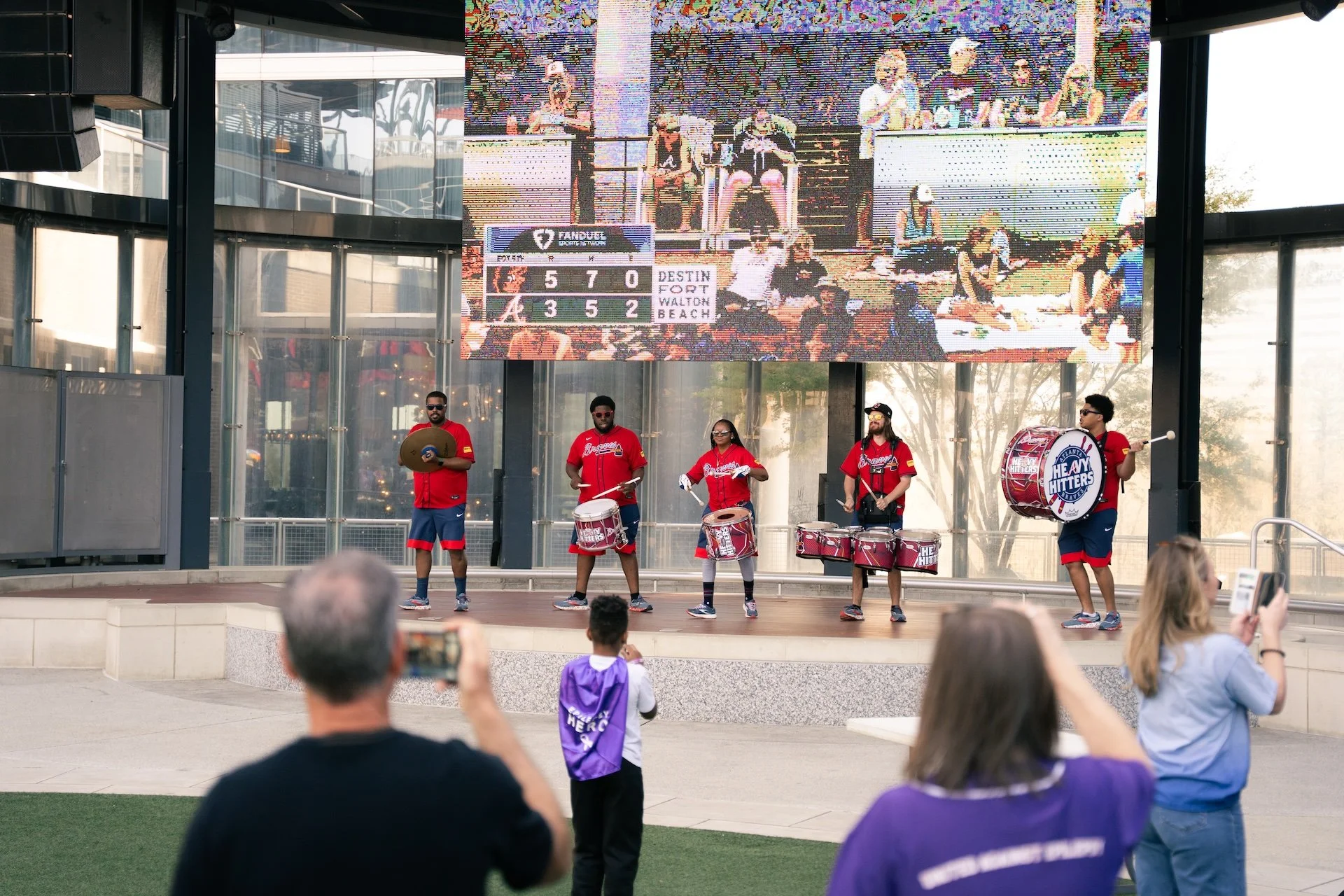 A band of five musicians, dressed in red and blue sports jerseys, performs on an outdoor stage with a large digital screen behind them showing a sports broadcast. The audience, including a man and a woman taking photos and a child in a purple shirt, 