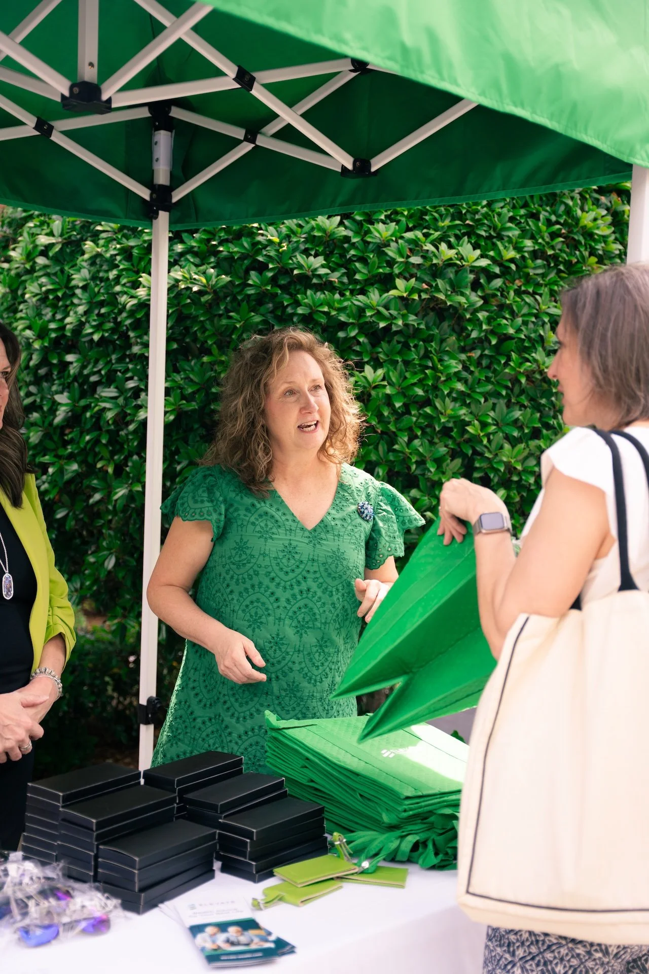 A woman in a green dress is speaking to a woman in a white top and carrying a white bag at an outdoor event under a green canopy. There are green tote bags, black boxes, and informational brochures on the table in front of them.