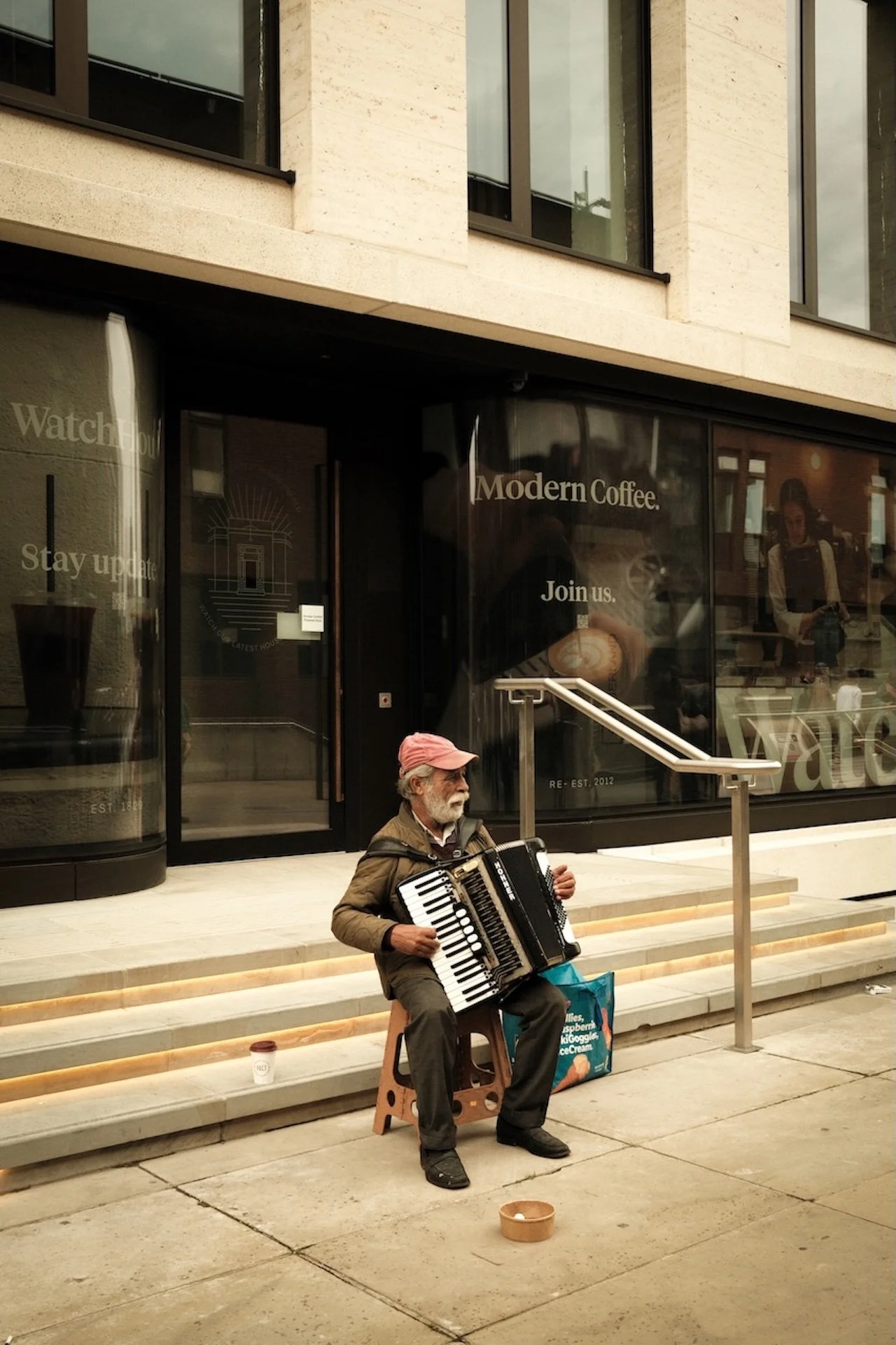 An elderly man with a white beard and wearing a red cap sits on a stool, playing an accordion on the sidewalk in front of a modern coffee shop.