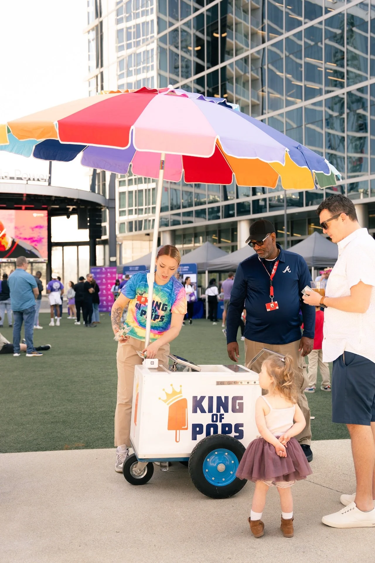 Girl selling popsicles from a cart labeled 'King of Pops' under a colorful umbrella at an outdoor event with people in the background.