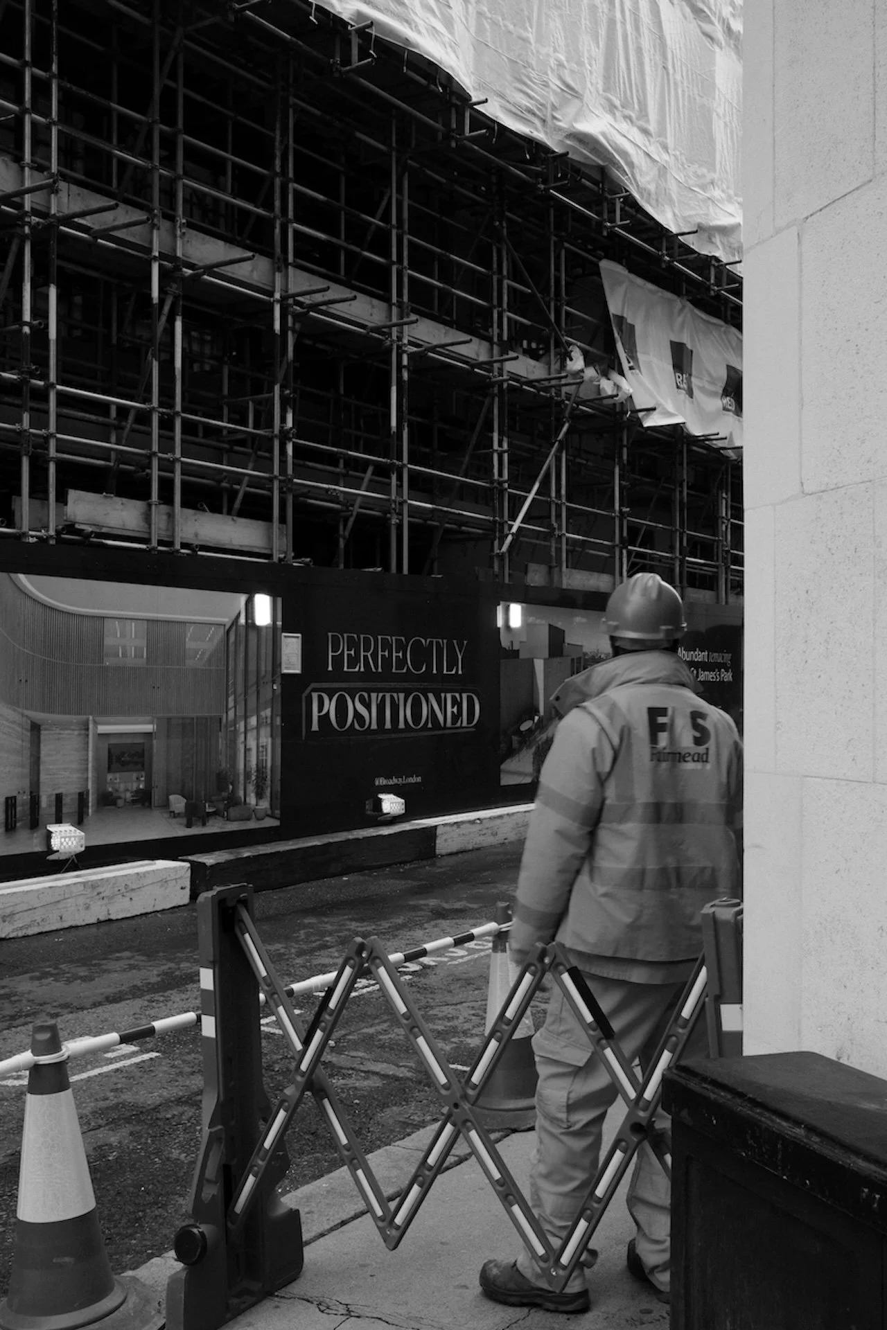 Construction worker in a helmet and vest standing behind safety barriers on a city street, with scaffolding and a sign that says 'Perfectly Positioned' in the background.