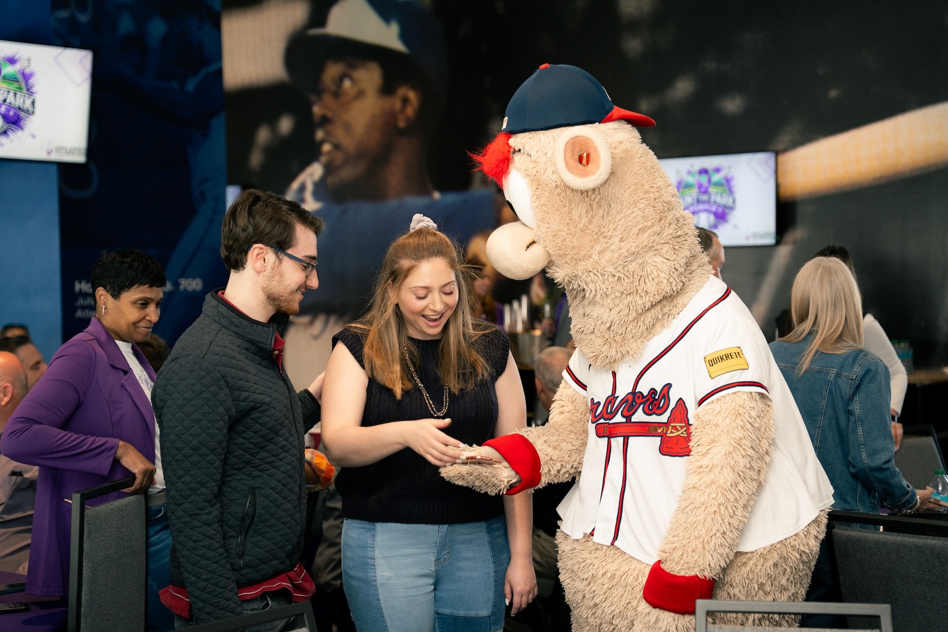 A woman smiling as she is shown something on her phone by a mascot wearing a baseball jersey and cap at a baseball-themed event, with people in the background