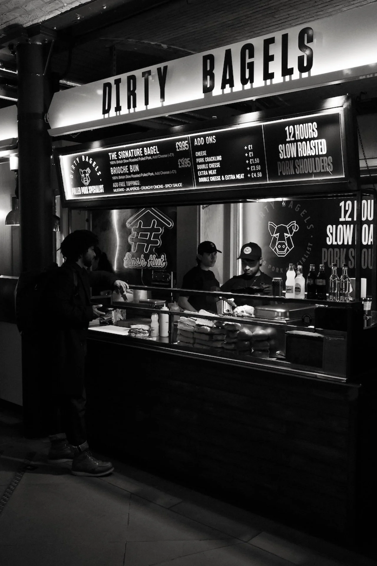 A food stall named 'Dirty Bagels' preparing sandwiches with customers in line. The stall has menus displaying bagel options, add-ons, and prices, with one customer waiting at the counter.