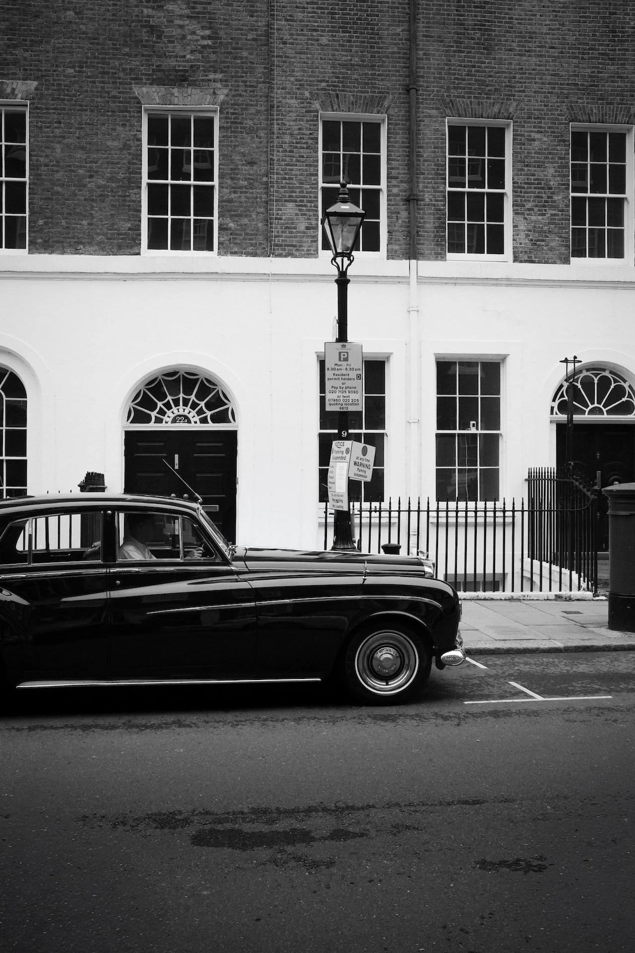 Black vintage car parked on the street in front of brick and white buildings with tall windows, a lamppost with parking signs, and a trash can.