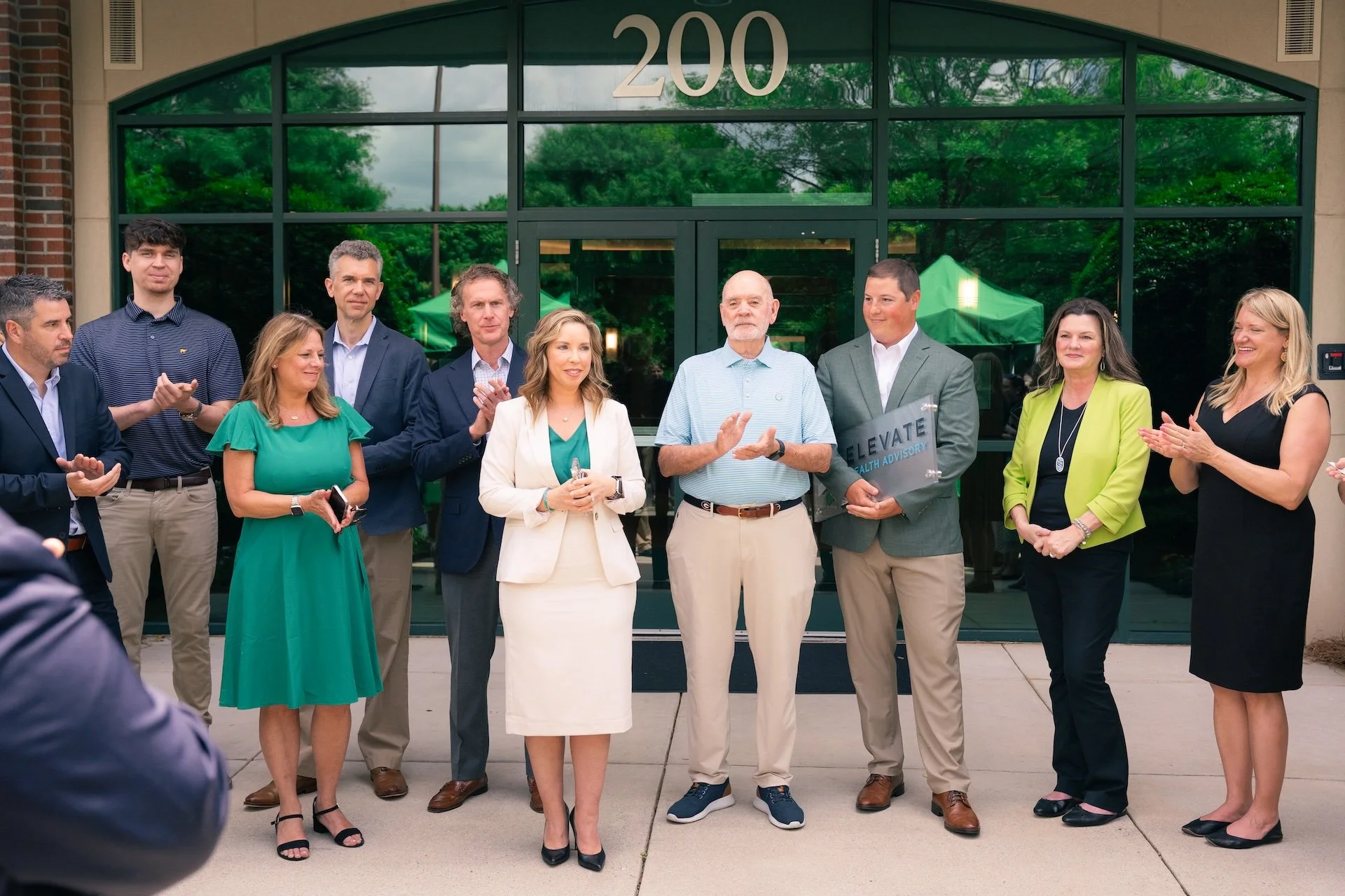 Group of professionally dressed people standing outside a building with the number 200 above the entrance, clapping and smiling during an event or celebration.