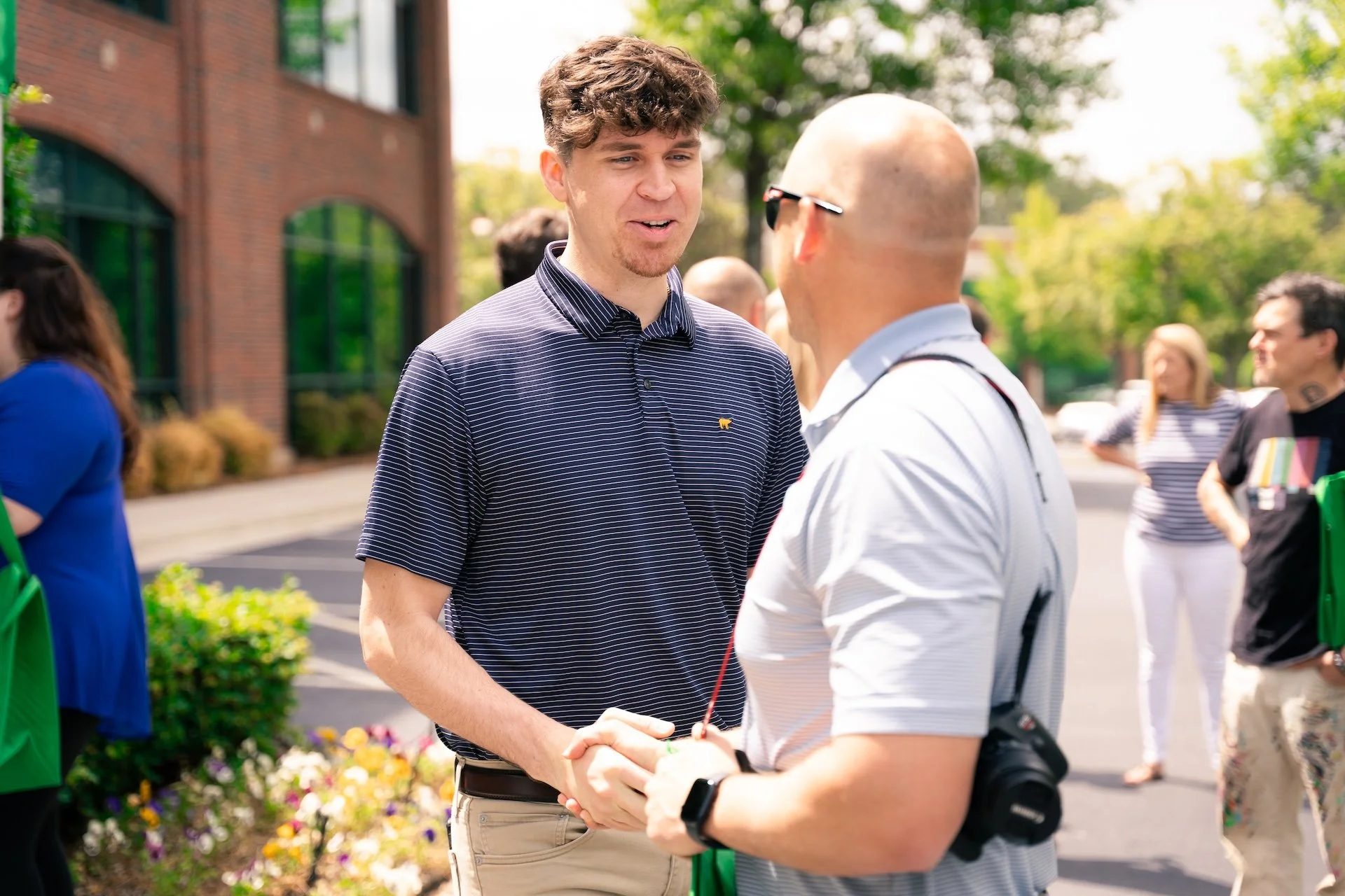 Two men shaking hands outdoors during a daytime event, with other people in the background near a brick building and greenery.