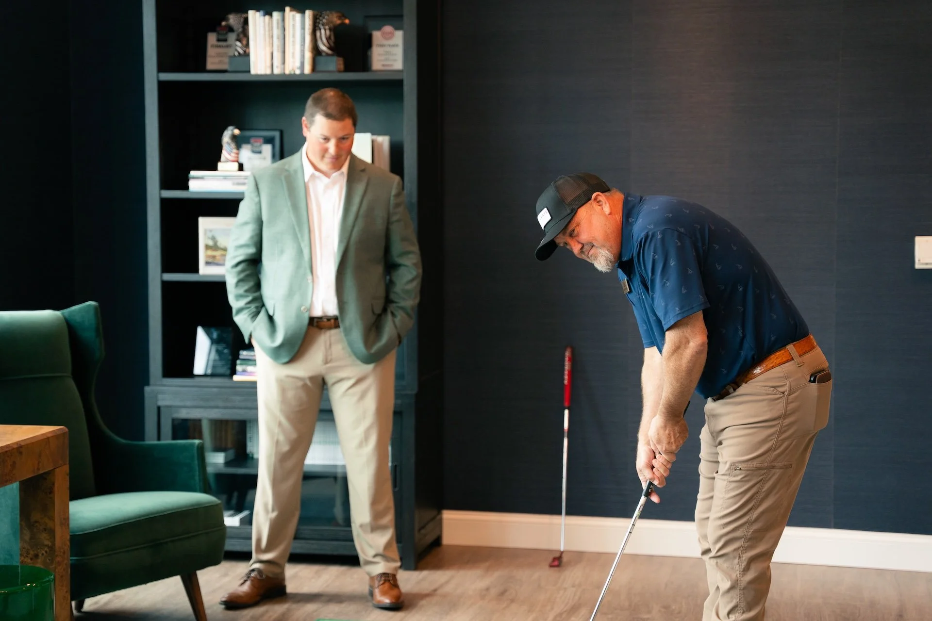 A man in a green blazer and beige pants stands observing another man in a blue shirt and tan pants playing golf indoors.