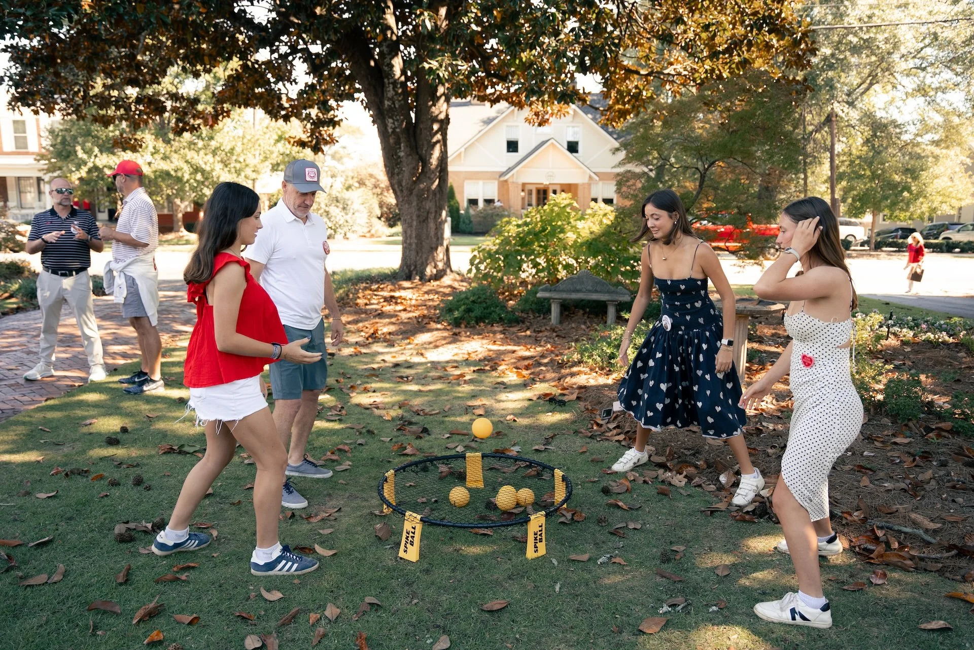 People playing a game with a yellow ball outdoors during daytime, with fallen leaves and trees in the background.