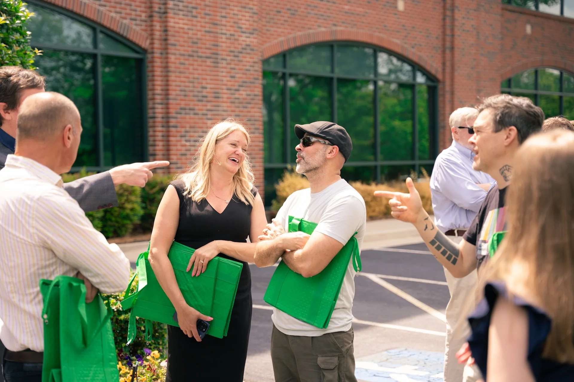 Group of people outside, talking and laughing, holding green folders or bags, in front of a brick building with large windows.