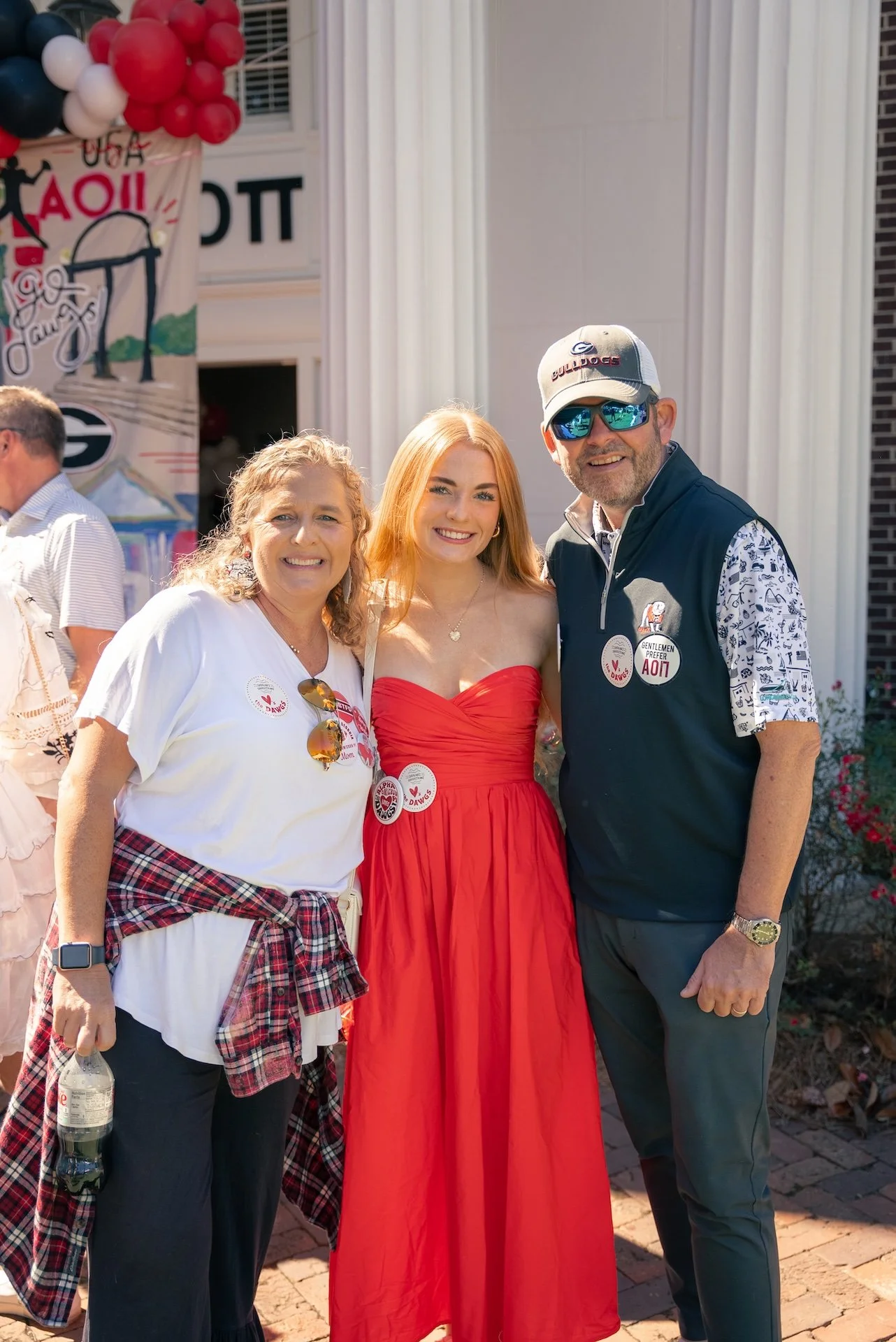 Three people smiling in front of a building with patriotic decorations. Two women and one man, all wearing badges.