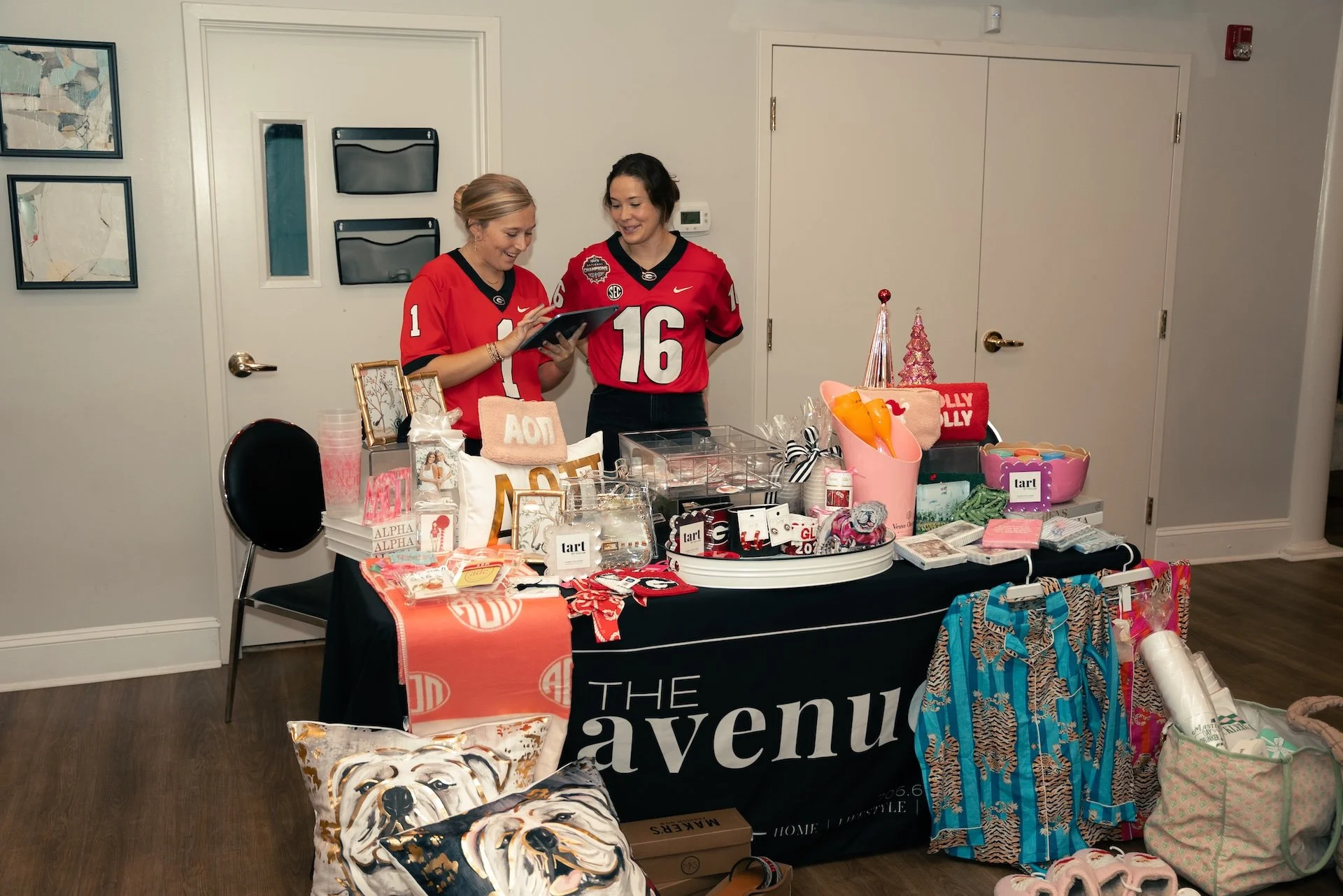Two women in red football jerseys standing behind a table with gifts and decorations, at a house party or celebration.