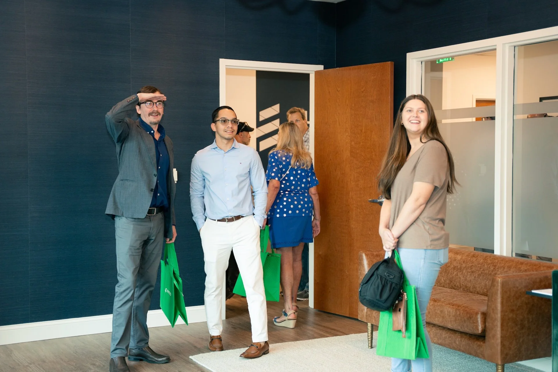 Group of people standing inside an office, some holding green tote bags, with one woman smiling at the camera near a brown couch.