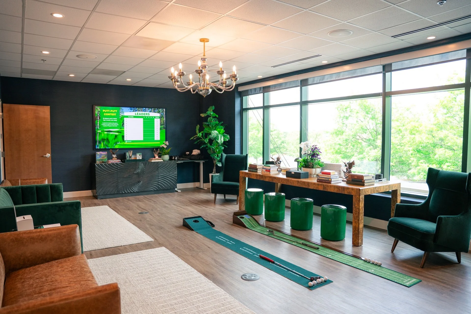 A spacious room with large windows showing green trees outside, decorated with a chandelier, potted plants, and various pieces of furniture, including a long wooden table with stacks of books, green stools, and a seating area with chairs and a rug.