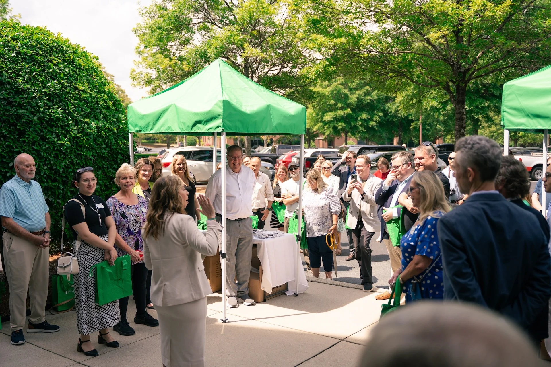 A woman speaks to a group of people gathered outdoors under green canopy tents, with everyone listening and smiling, during a daytime event on a sunny day.