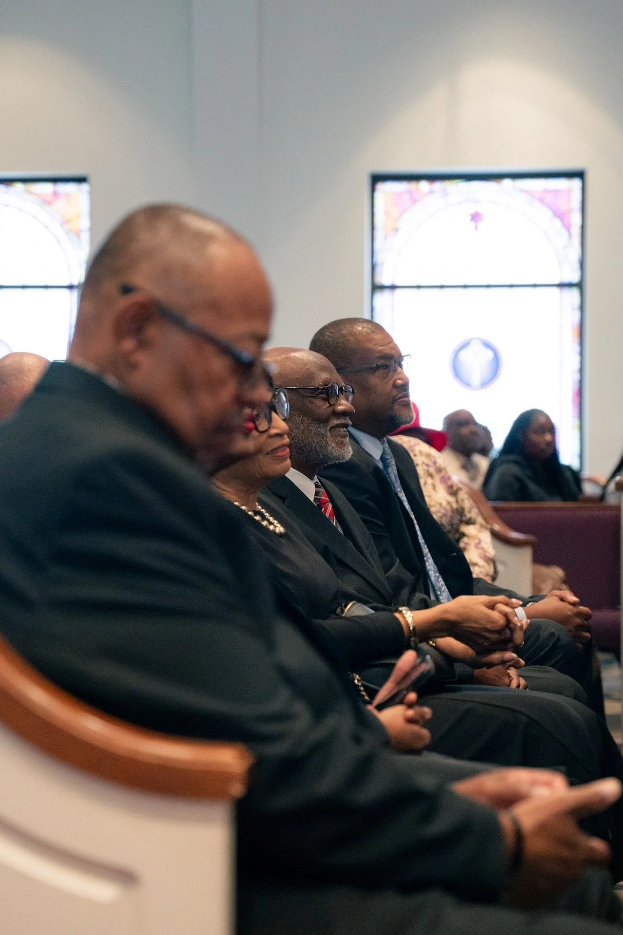 Group of people sitting in pews inside a church, with stained glass windows in the background, attending a formal service or event.
