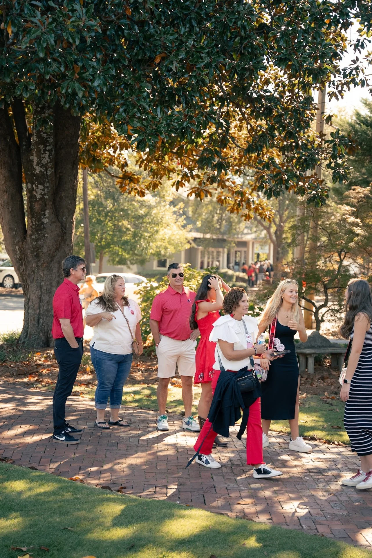 A group of diverse people standing outdoors on a brick sidewalk in front of a large tree, engaging in conversation and smiling, with a house and cars in the background.