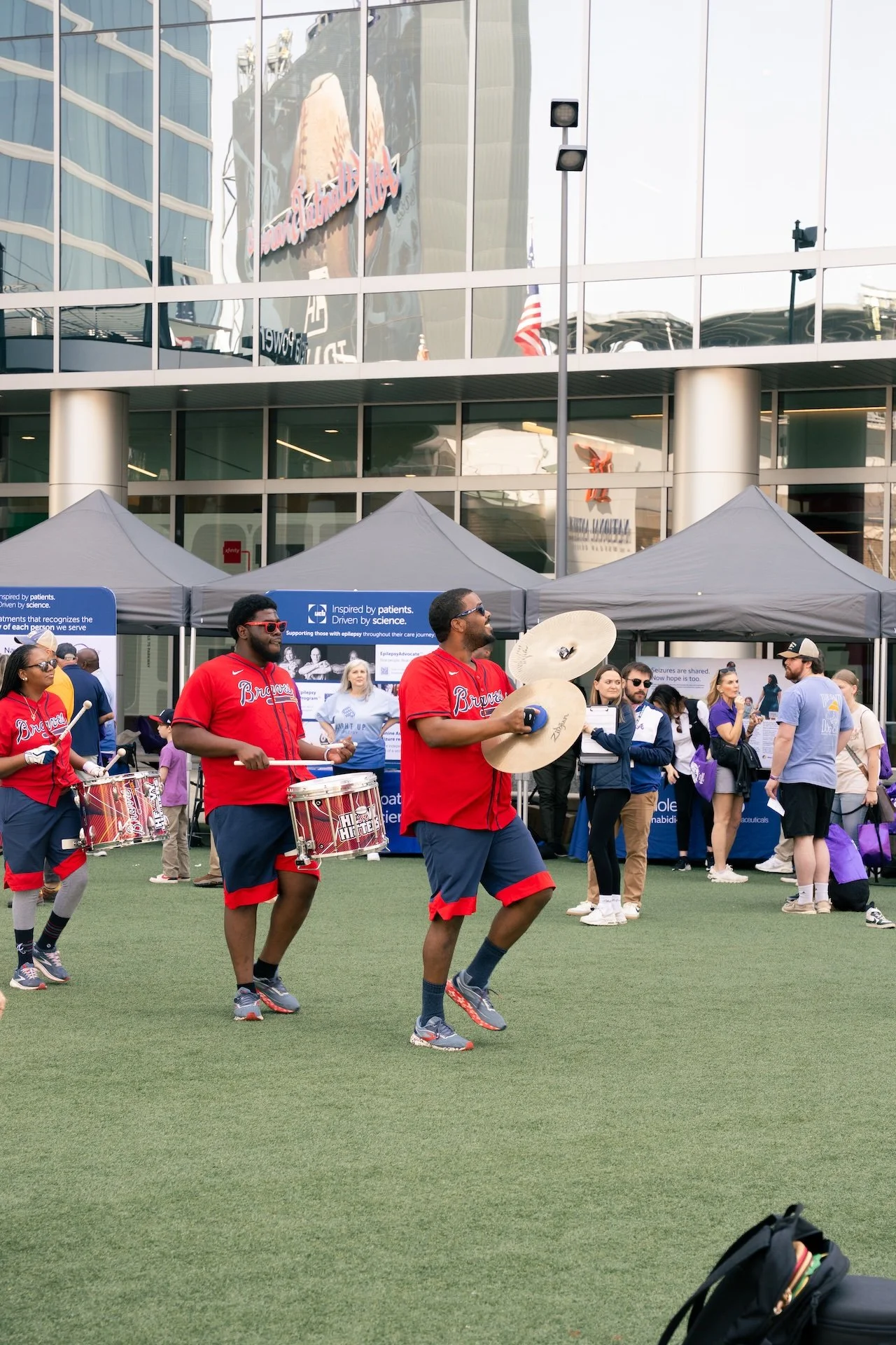 Musicians play drums and cymbals at an outdoor event with tents and groups of people, some wearing Atlanta Braves jerseys, in front of a modern glass building.