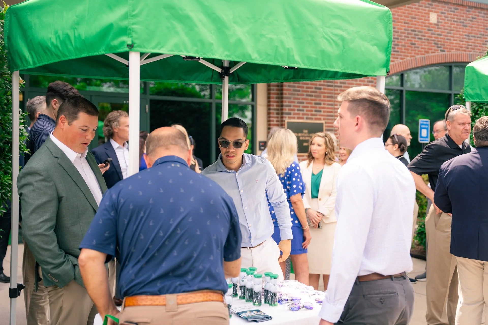 People gathered outdoors under green tents at a professional or networking event on a sunny day.