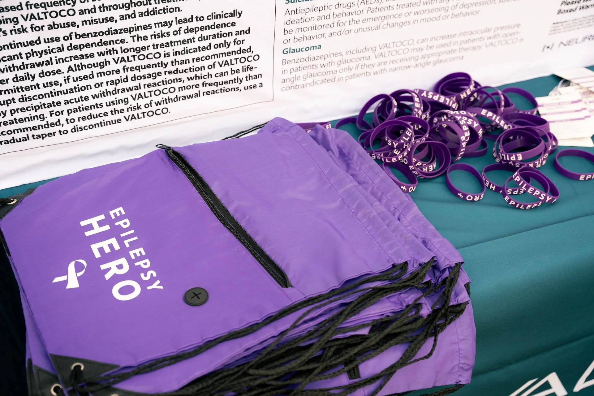 A purple tote bag labeled 'Epilepsy Hero' and purple silicone bracelets with 'Epilepsy Hero' written on them, displayed on a teal table near an informational poster.