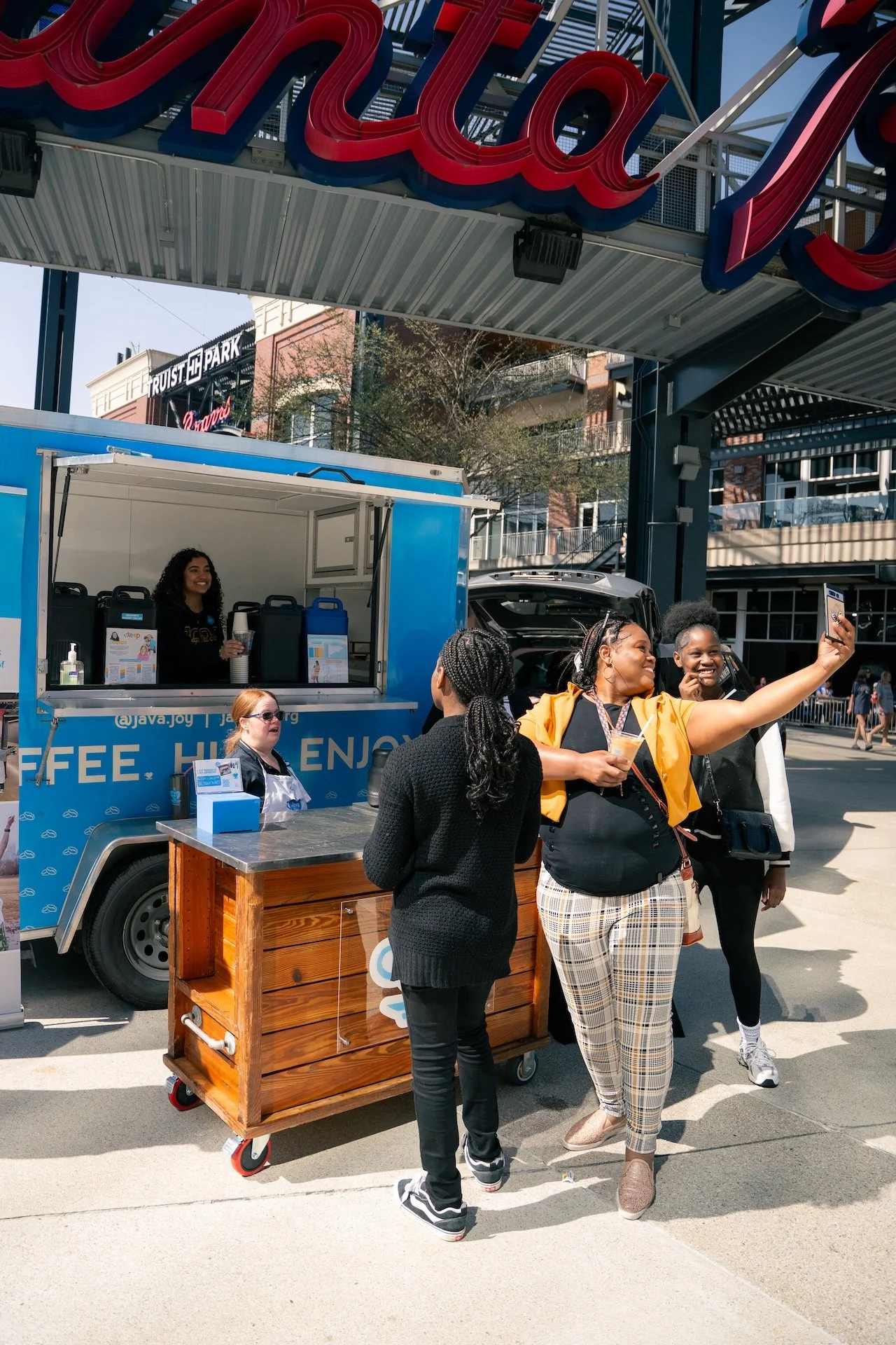 A group of four women standing outside a coffee truck. One woman is taking a selfie with her phone. The coffee truck has a smiling barista inside, and there is a small wooden cart nearby. In the background, there is a large sign with the word 'Russ'.
