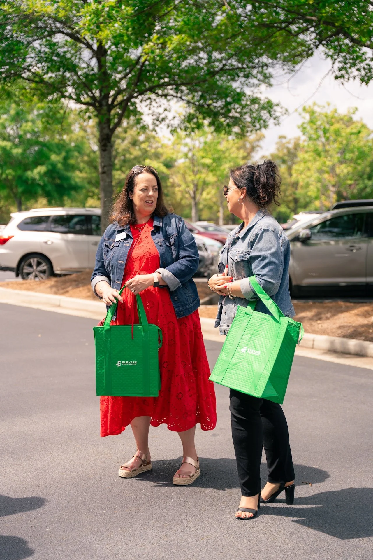 Two women talking outdoors in a parking lot, carrying green reusable shopping bags, with trees and parked cars in the background.