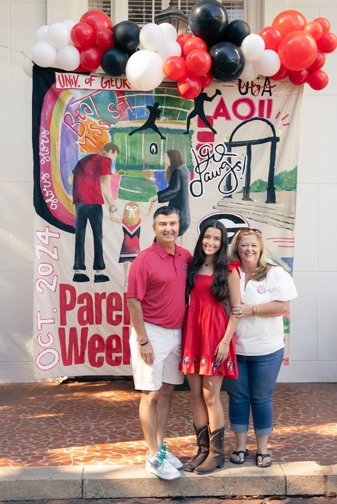 Three people standing in front of a large colorful banner and balloons during a university event. The banner features drawings of students and sports, with the text 'Parent Week' and date 'October 24, 2023'.