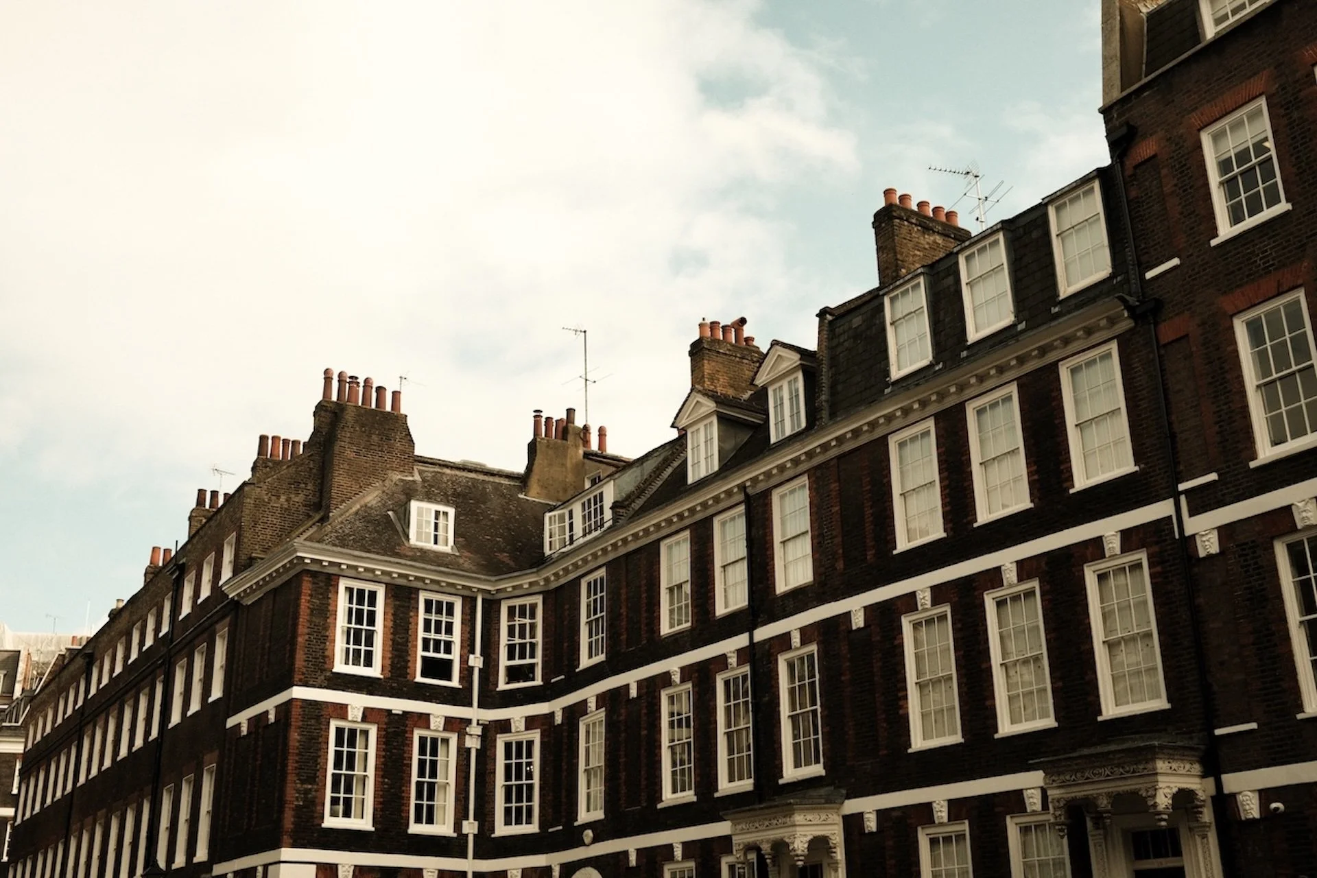 Multiple old brick buildings with white-framed windows and rooftops with chimneys under a partly cloudy sky.