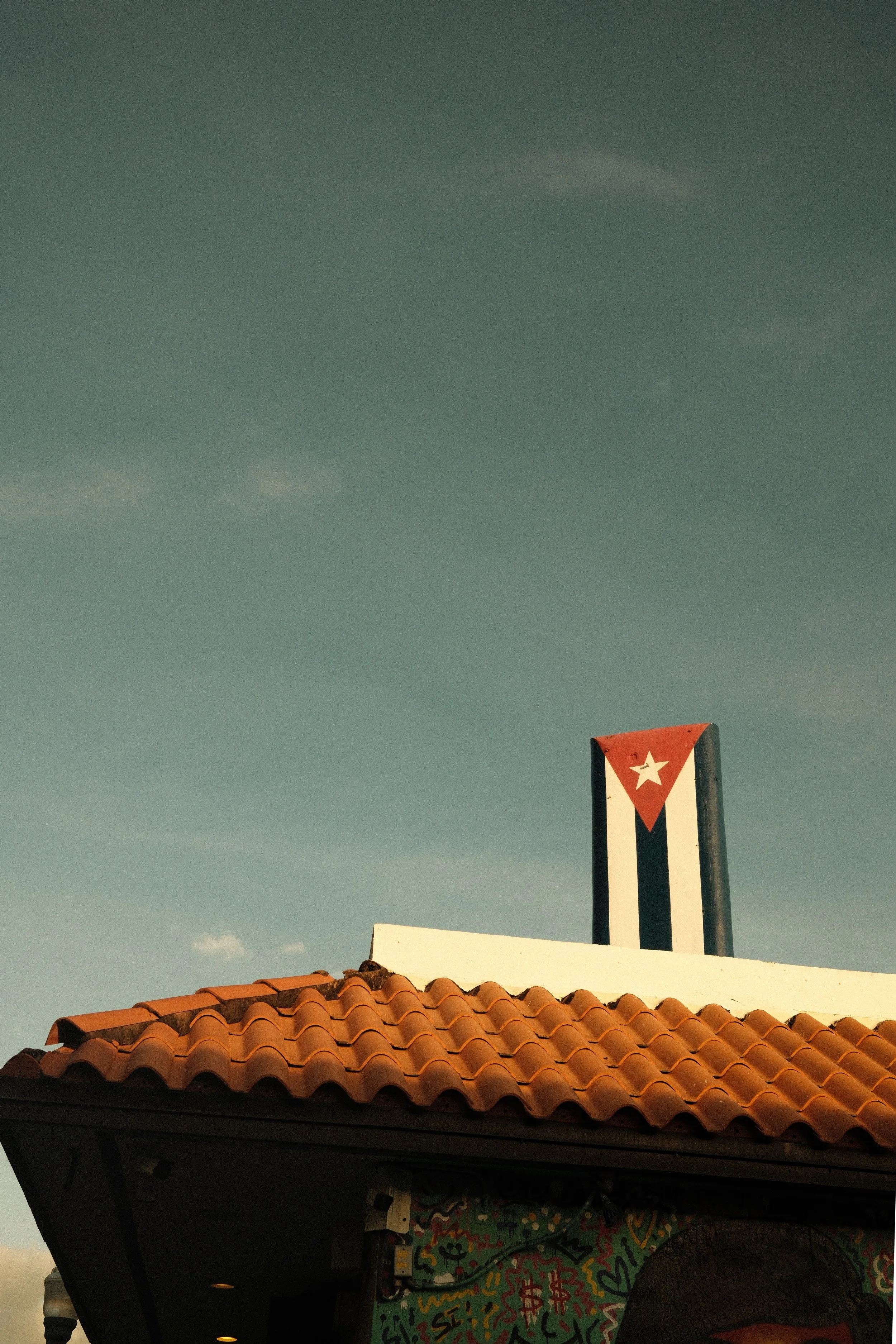 Part of a building with a red tiled roof, and a sign with a Cuban flag, featuring a white star and red, white, and blue stripes, against a clear sky.