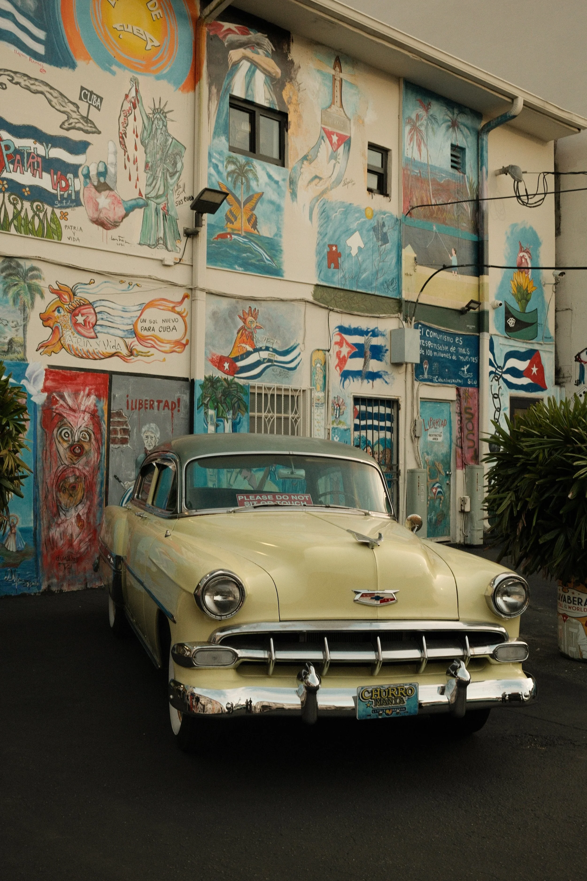 A vintage yellow Chevrolet car parked in front of a colorful mural wall in Cuba, featuring various images and symbols related to Cuba and revolution.