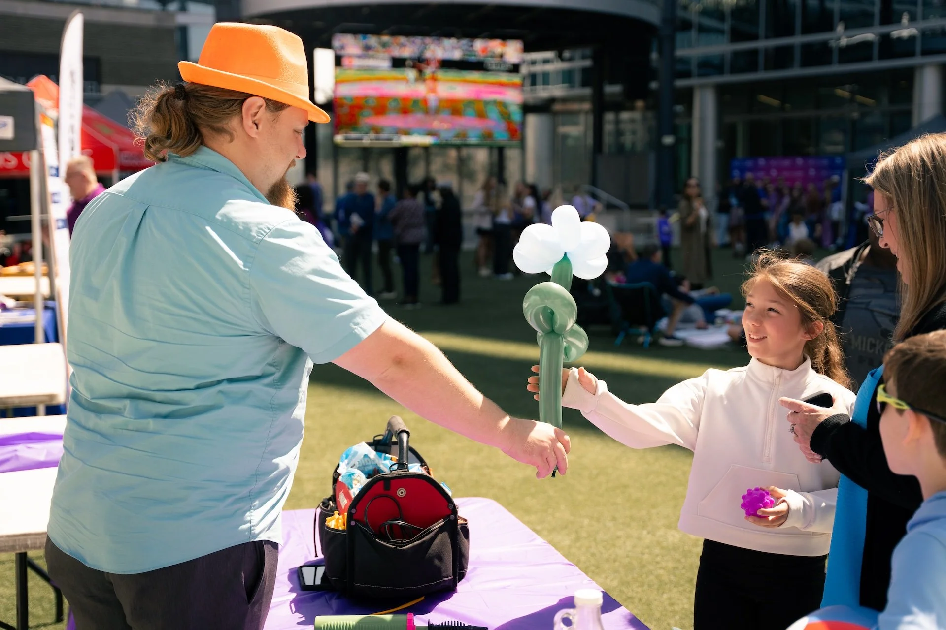 A man in an orange hat and light blue shirt hands a balloon flower to a smiling girl in a white jacket at an outdoor event. Other children and adults are visible in the background.