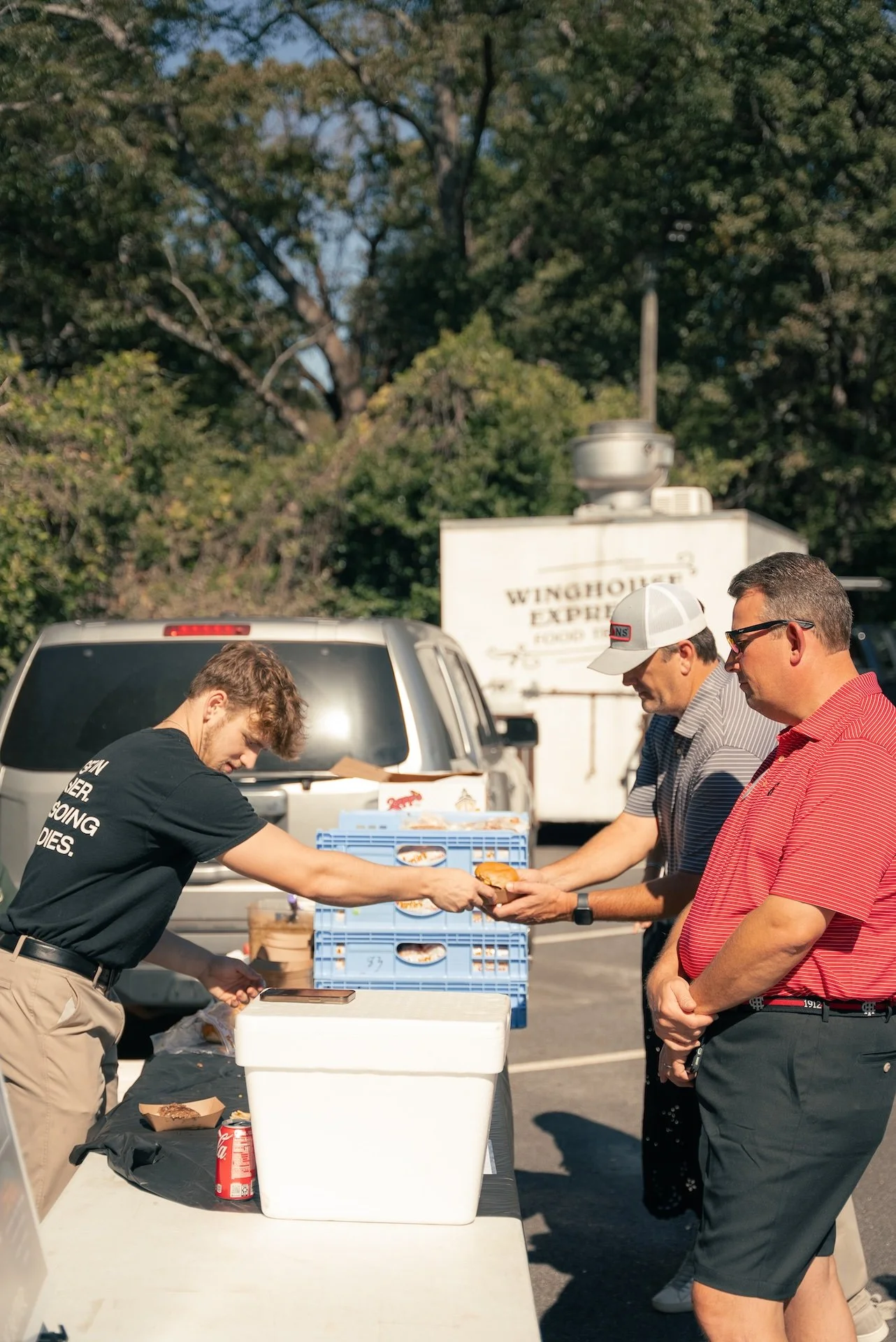 Three men exchanging a hot dog at an outdoor food stand in a parking lot with trees in the background.