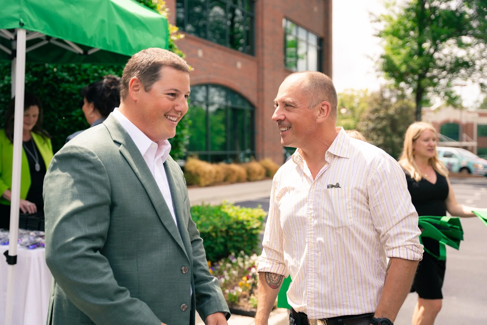 Two men are smiling and talking outdoors at a daytime event, with people and a building in the background.