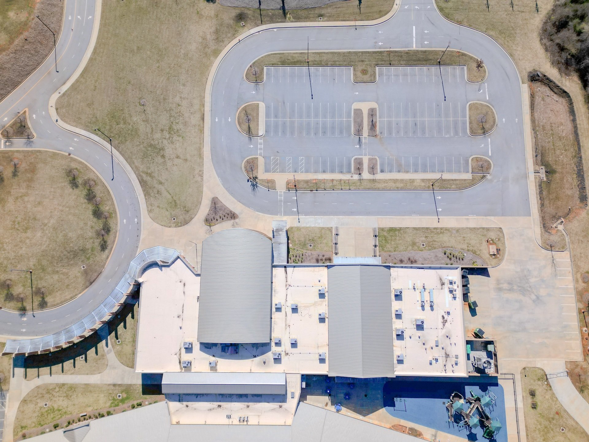 An aerial view of a large parking lot with several lined parking spaces, surrounded by green grassy areas, trees, pathways, and playground equipment.