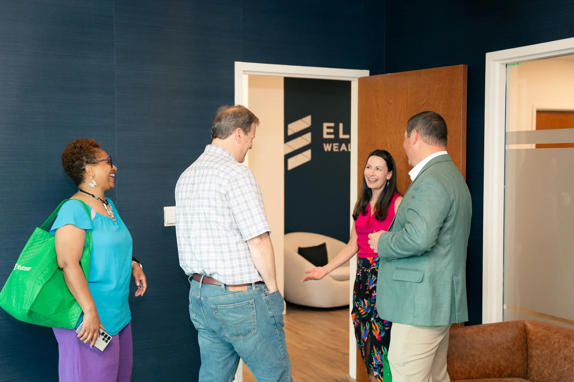Four people are standing and chatting in an office, laughing and smiling. The group consists of two men and two women, one of whom is holding a smartphone. They are gathered near a door with a sign that reads 'ELITE WEALTH'. The office has navy blue 