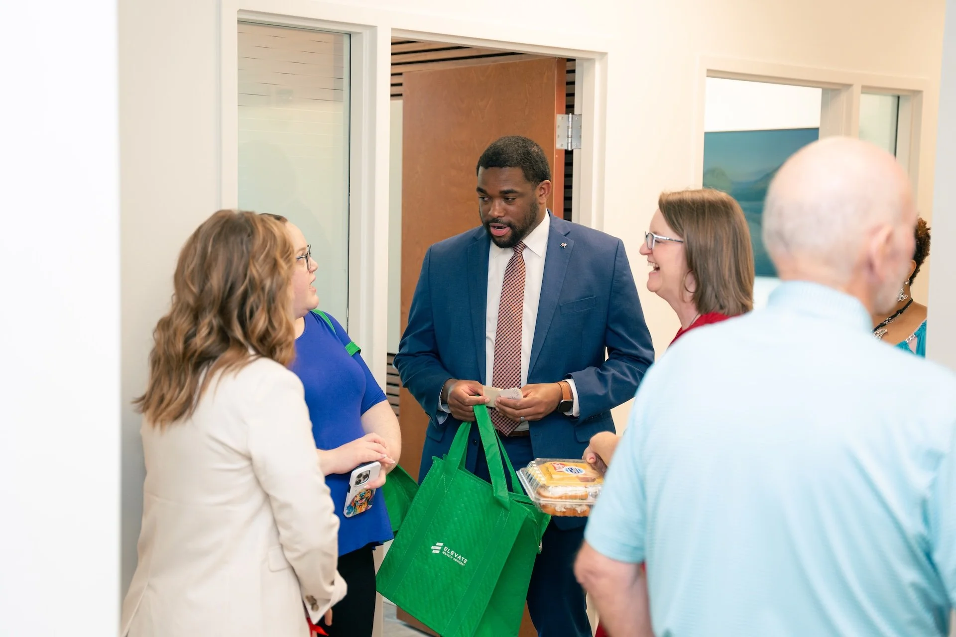 A man in a blue suit holding a green shopping bag and a food container, standing and talking to three women and a man at an indoor gathering.