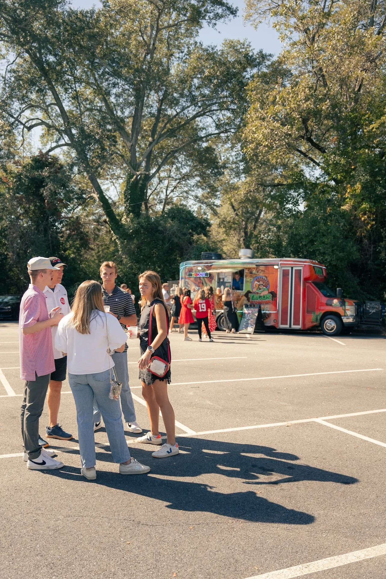 Group of people standing in a parking lot near a colorful taco food truck on a sunny day.