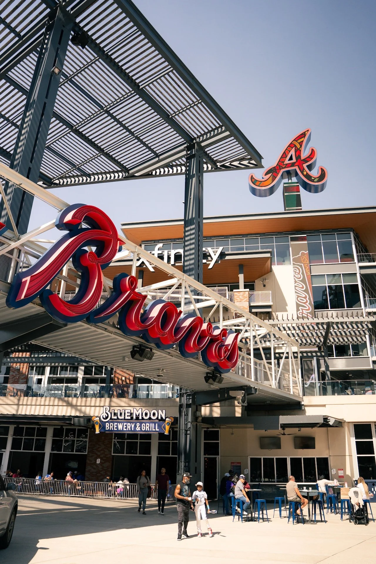 Exterior view of a shopping and dining complex with signs for Tijuana, Blue Moon Brewery & Grill, and an arcade. There are people walking and sitting outside under a clear sky.