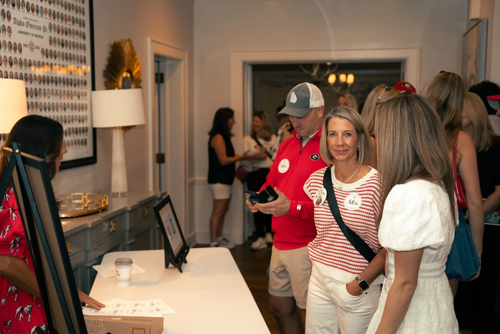 People gathered at an indoor event, engaging with a registration or check-in desk, with one woman smiling at the camera, and others focused on the desk or talking in the background.