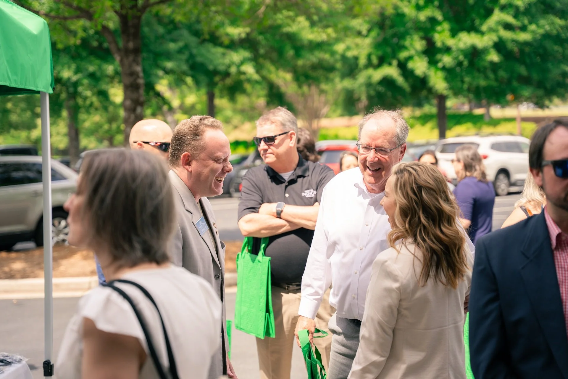 People smiling and chatting outdoors at a sunny event, with parked cars in the background.