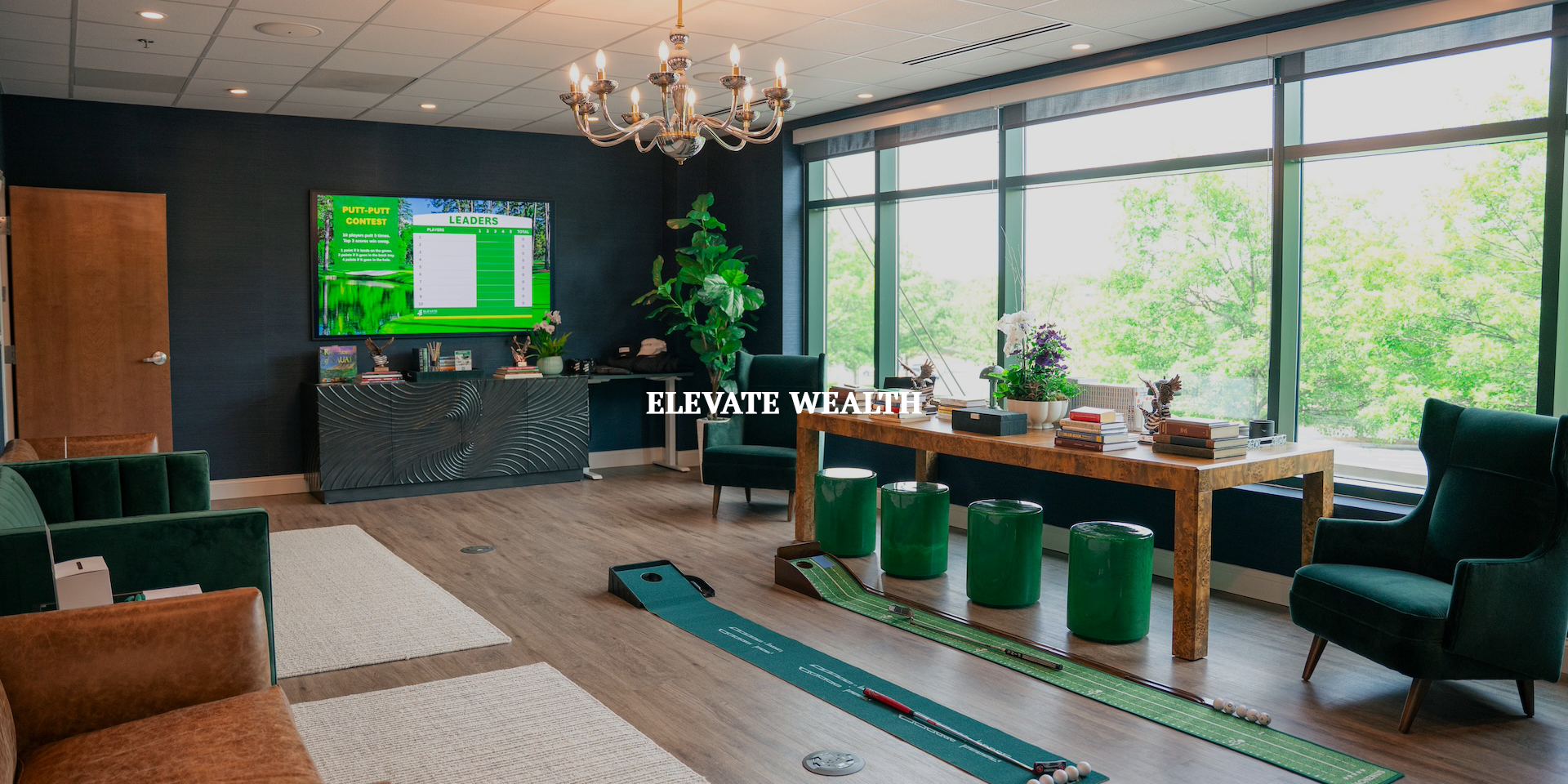 Interior of a modern office with a large window showing green trees outside. There are green and brown chairs, a wooden table with golf putting mats and golf balls, books and plants on a side table, a large TV on a dark wall displaying a golf leaderboard, a chandelier hanging from the ceiling, and decorative items on a console.