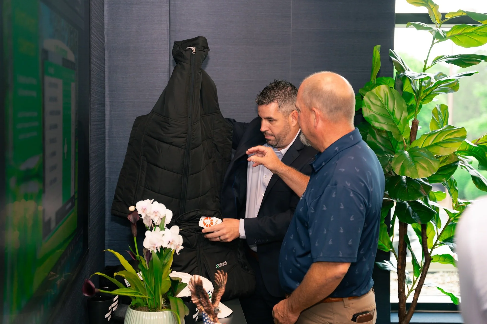 Two men are standing near a wall with a hanging black jacket. One man is holding a small box with an orange logo, and the other is pointing at it. There are flower arrangements and potted plants nearby, with a window in the background showing greener