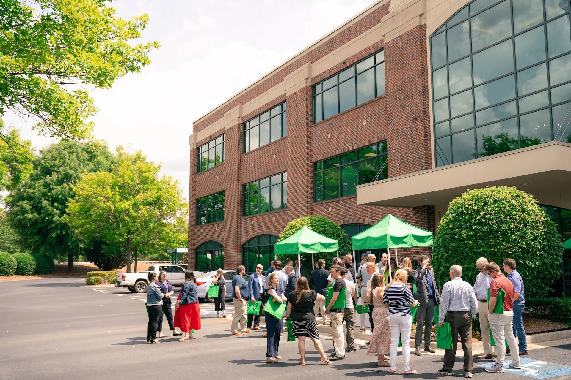 People gathered outside a brick office building under green tents, carrying green tote bags, engaging in conversation, with trees and parked cars nearby.